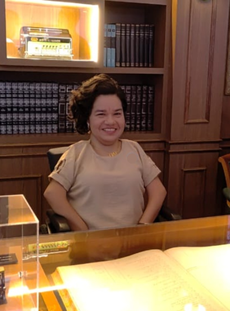 Foto da autora do blog, Sandra Menezes, que está sentada a uma mesa com um livro Amarelo aberto atrás de si uma estante de livros antiga
a mesma está sentada na cadeira de rodas olhando para câmera sorrindo vestindo uma blusa cor de café com leite e ela tem os cabelos pretos curtos ondulados